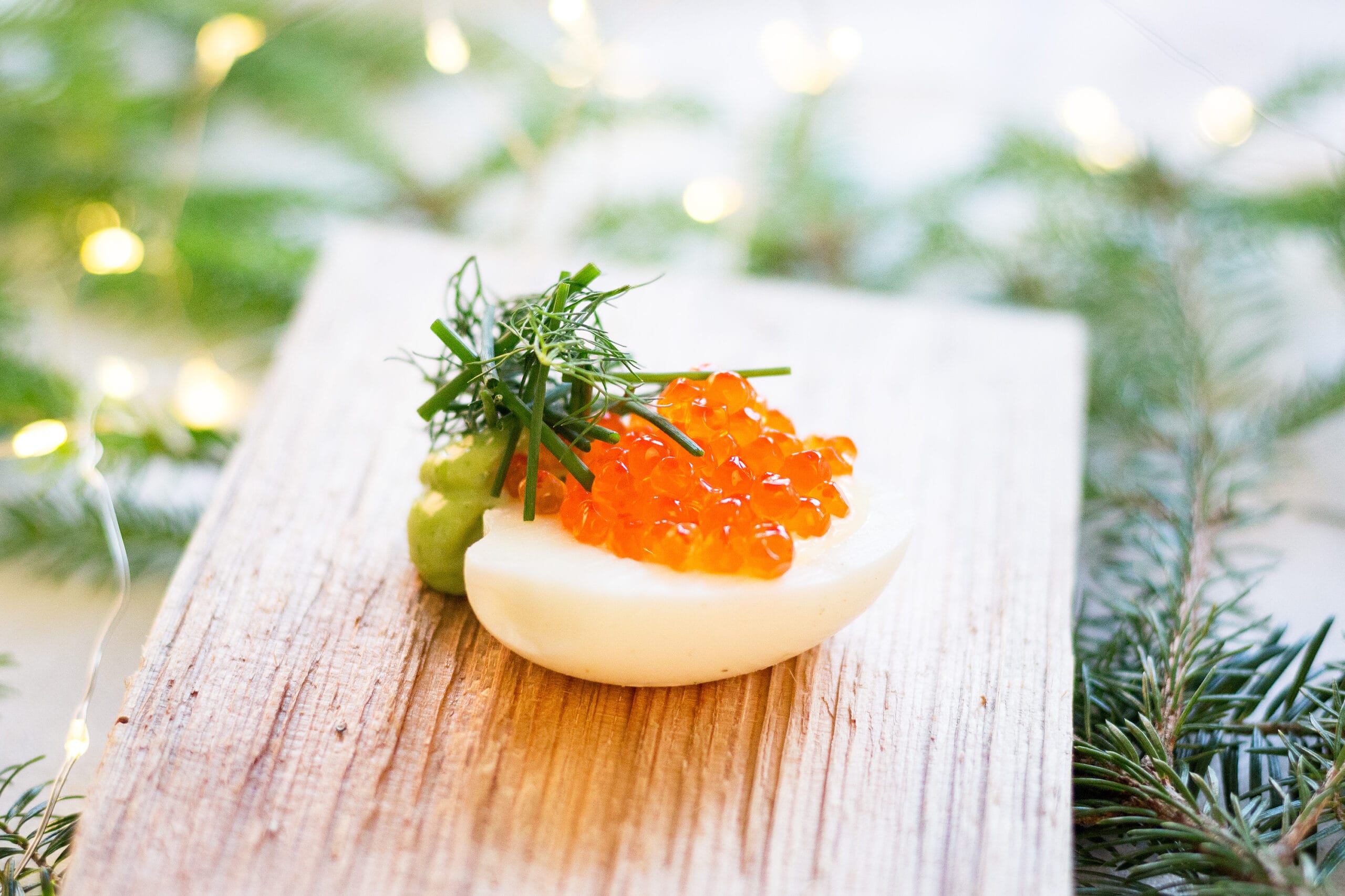 Garnerad ägghalva En skiva ägg toppad med orange fiskrom, färska örter och grön garnityr, serverad på en rustik träbräda med suddiga ljus och vintergröna grenar i bakgrunden.
