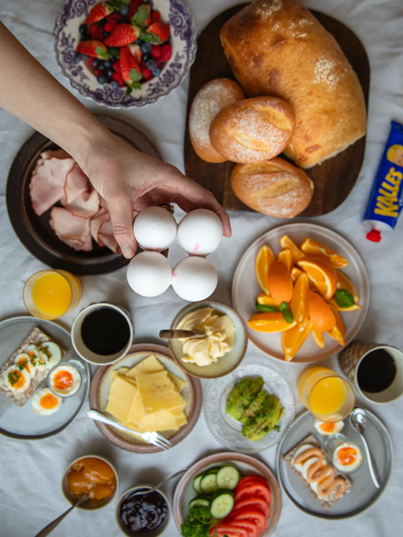 Ett bord dukat med olika frukosträtter: bröd, frallor, ost, skinka, skivade grönsaker, frukt, apelsinskivor, sylt, avokado, ägg, juice, kaffe och en hand som håller i fyra vita ägg ovanför upplägget.