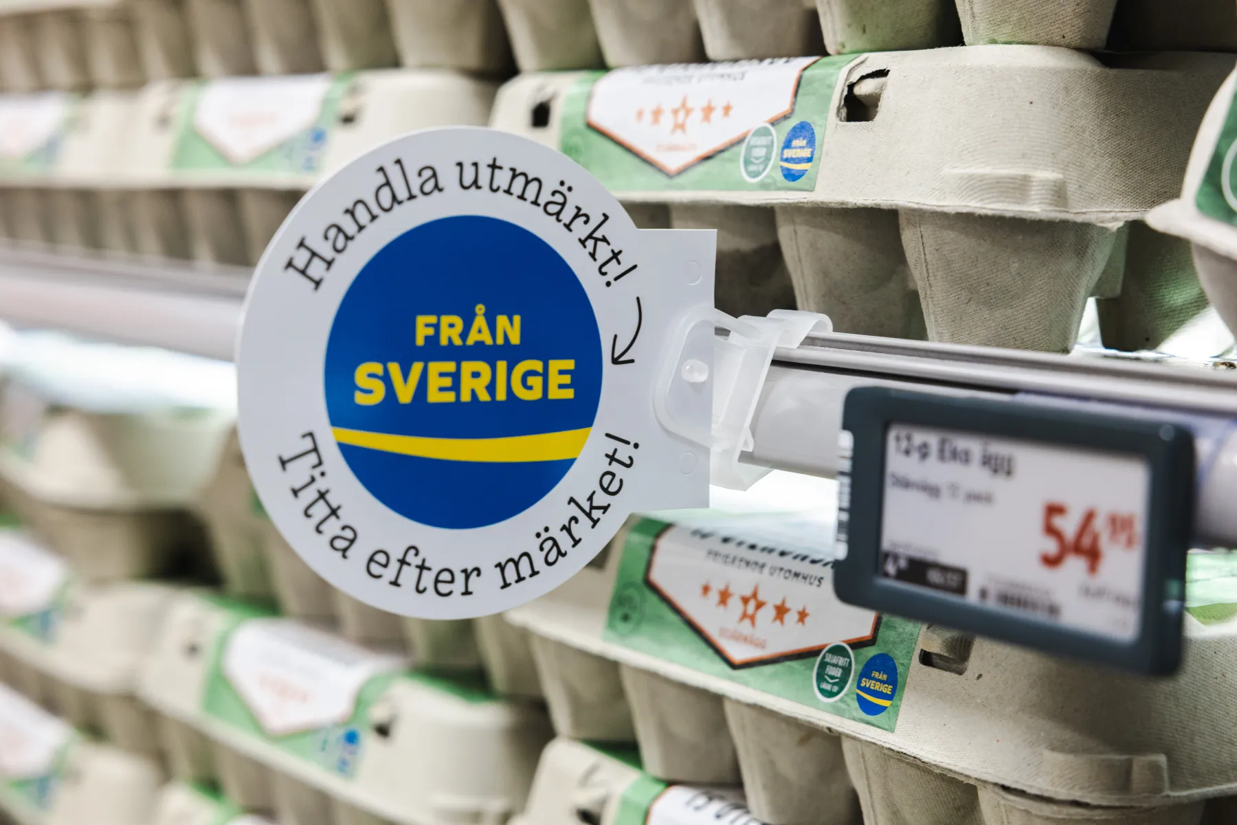En "Från Sverige"-skylt är fäst på en hylla i ett snabbköp som är full av äggkartonger. En digital prislapp visar 54,95 kronor. Skylten uppmuntrar till att köpa svenska produkter med svensk text.