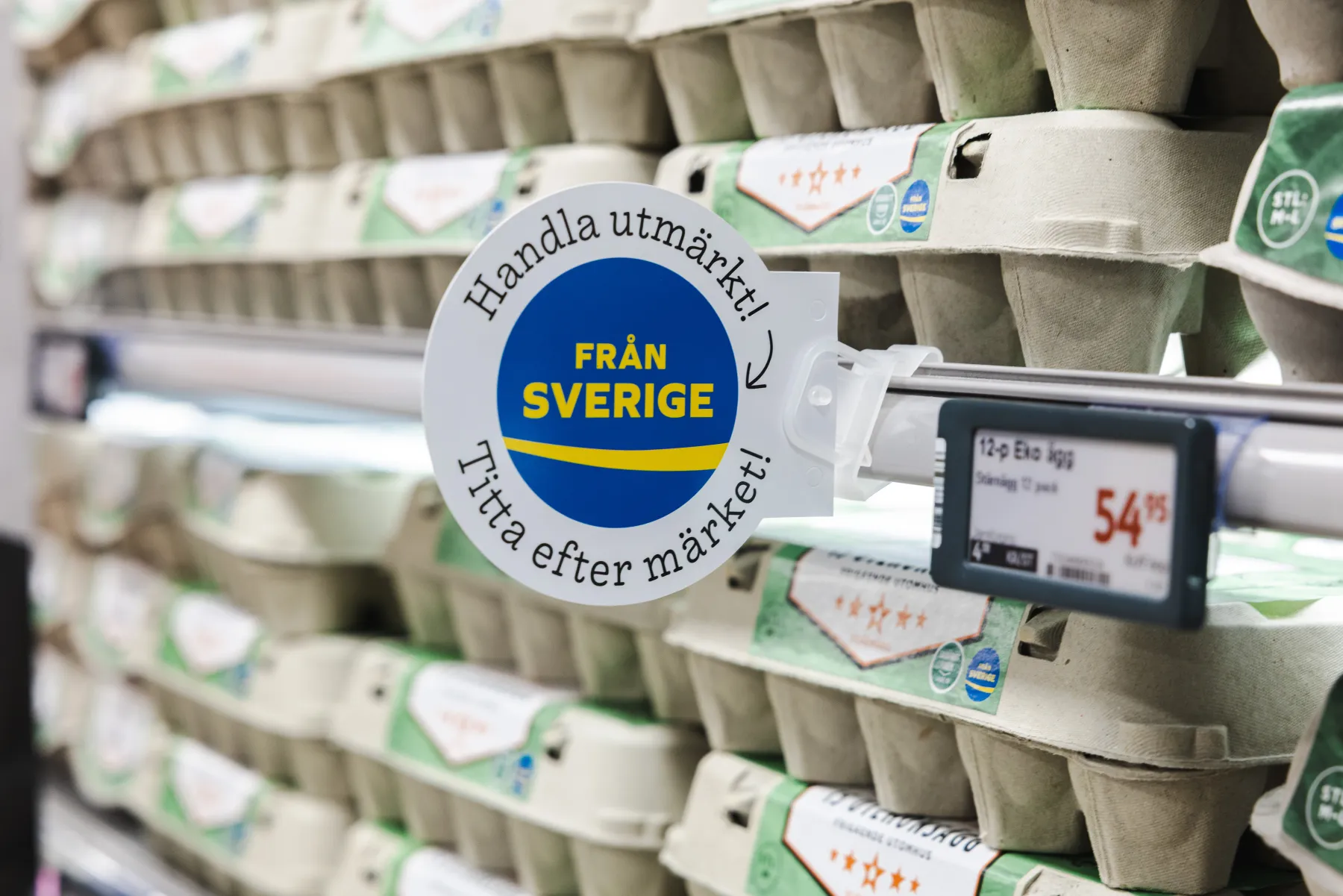 En butikshylla fylld med äggkartonger. En rund skylt på svenska, med blå och gula färger, med texten "Från Sverige" uppmanar kunderna att leta efter svenska produkter. En digital prislapp visar 54,95 kr.