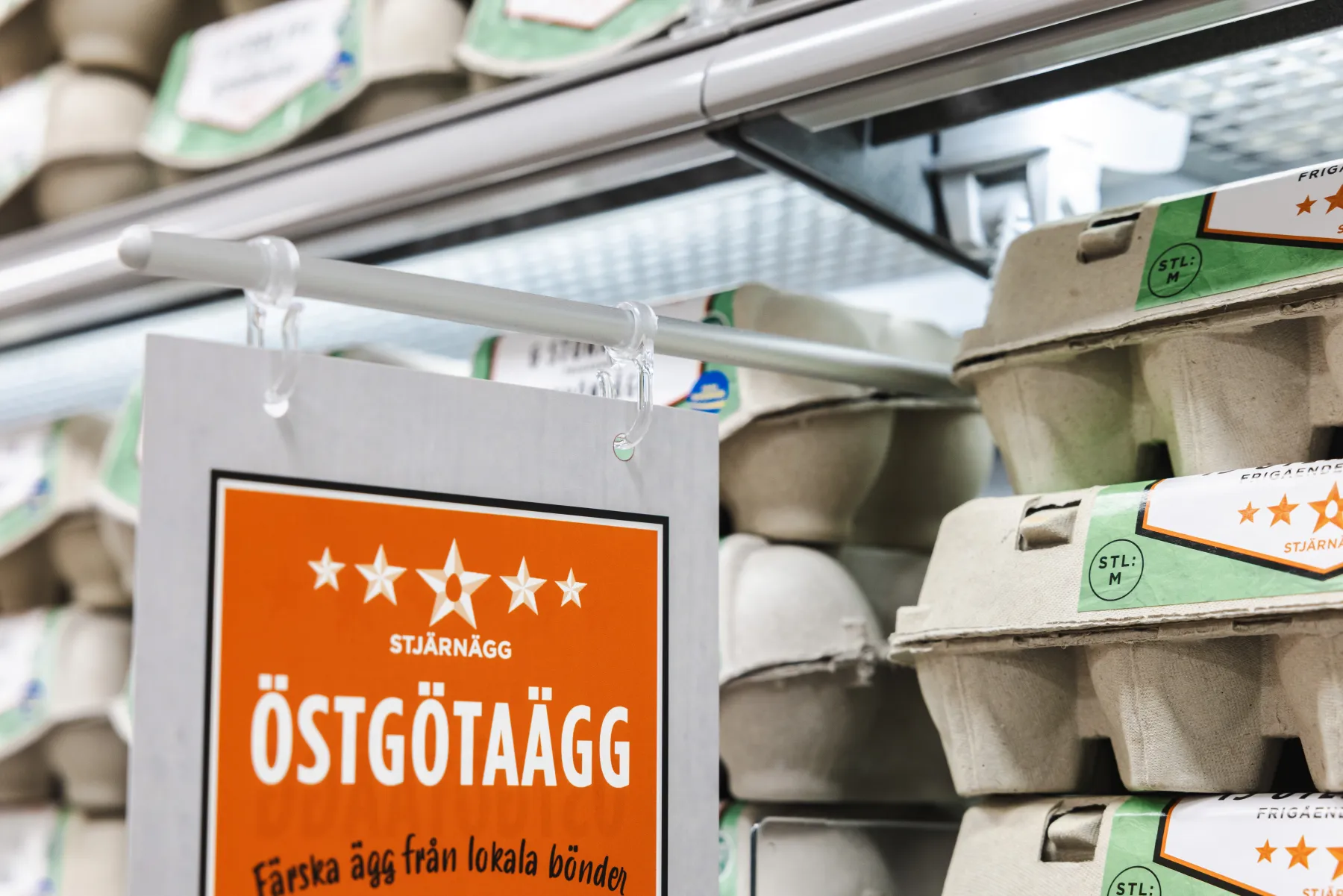 Äggkartonger staplade på en hylla i ett snabbköp bredvid en hängande orange skylt med texten "Östgötaägg", som gör reklam för färska lokala ägg på svenska.
