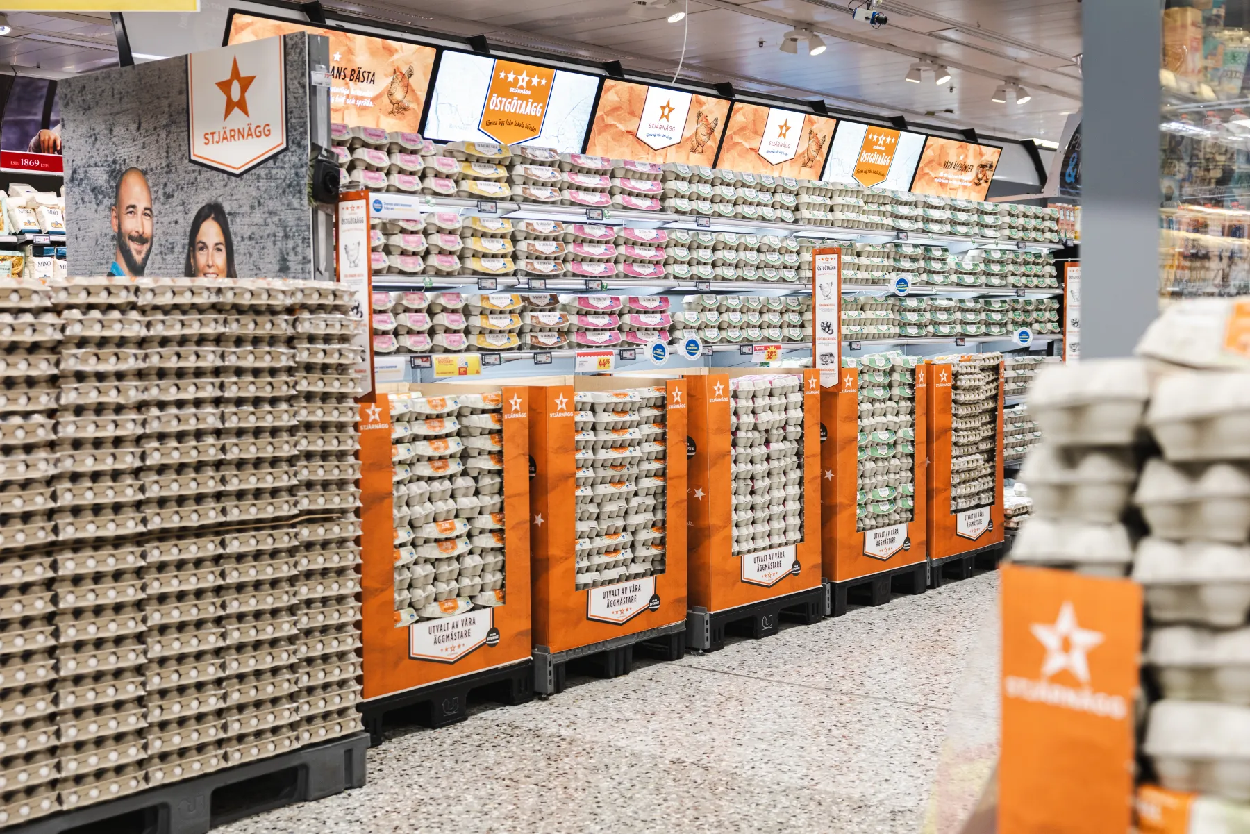 I en stormarknadsgång står staplar av äggkartonger på hyllor och i orangea fack. Ovanför hänger skyltar med stjärnor och förpackningarna är prydligt ordnade över hela avdelningen. Golvet är fläckigt och en del av en annan hylla syns till höger.