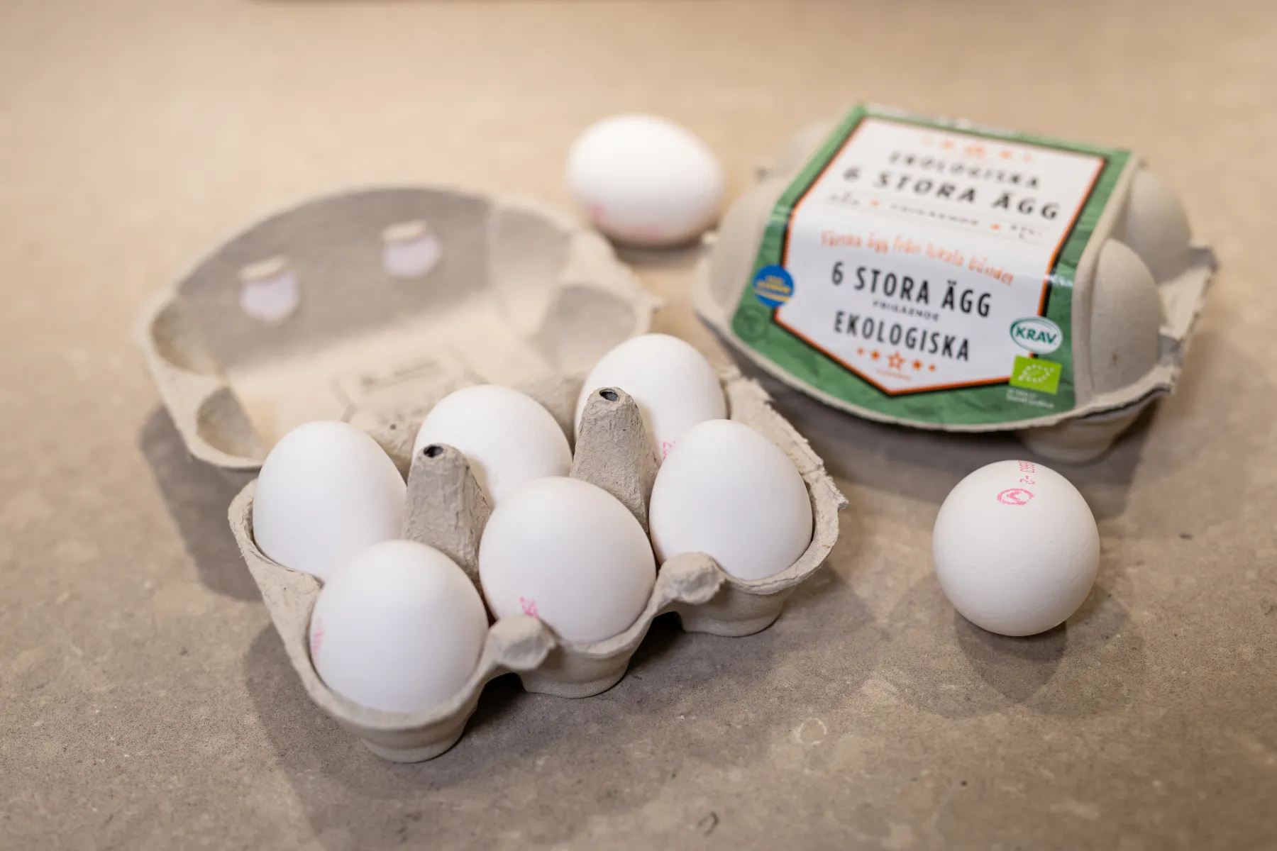 En kartong med sex vita ägg ligger öppen på en bänkskiva och ett ägg har tagits ut och placerats i närheten. En annan stängd kartong märkt "6 stora ägg ekologiska" finns i bakgrunden.