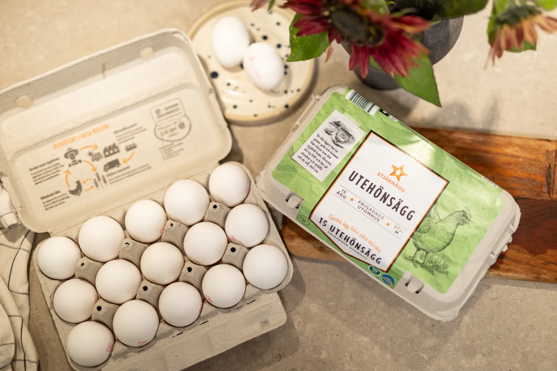 En kartong med vita ägg ligger öppen på en bänkskiva bredvid en stängd kartong märkt Utehönsägg med illustrationer av kycklingar. I närheten finns två lösa ägg och en vas med blommor.