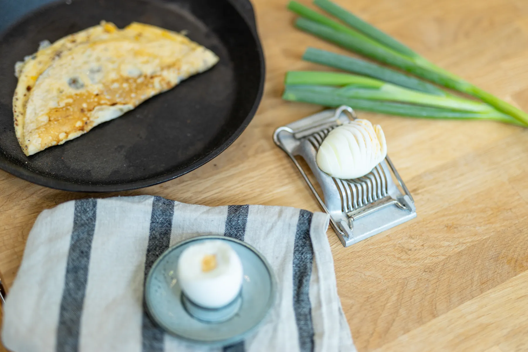 Ett skivat hårdkokt ägg ligger på en äggskivare bredvid salladslök, med en kokt omelett i en kastrull och ett mjukkokt ägg i en äggkopp på en randig duk, allt på en träyta.