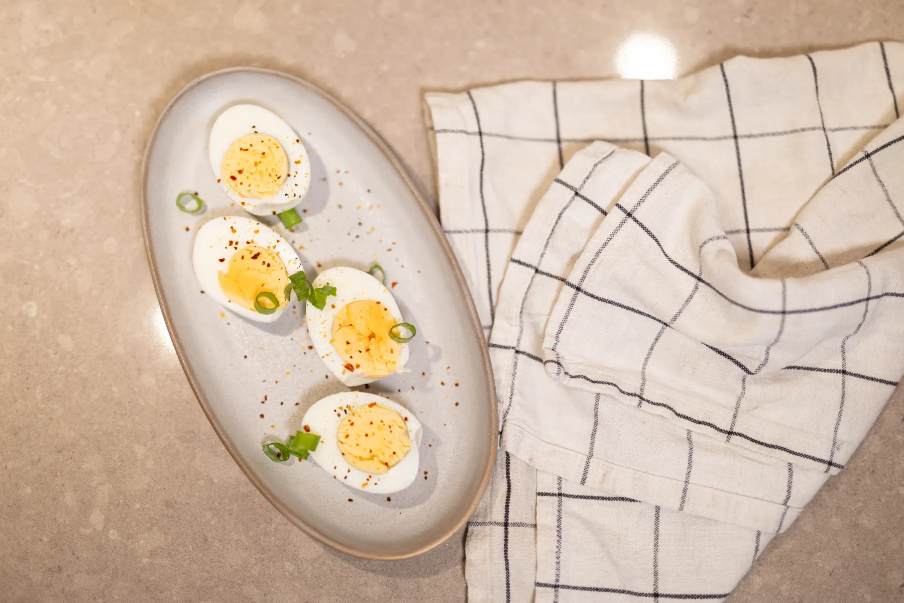 Fyra deviled eggs garnerade med salladslök och rödpepparflingor ligger på en oval tallrik bredvid en vit- och svartrutig kökshandduk på en beige bänkskiva.