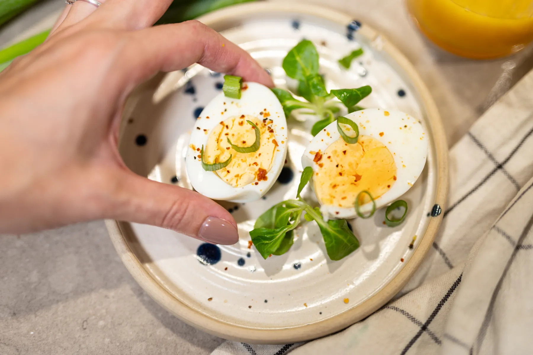 En hand håller i en halv hårdkokt ägg, garnerad med salladslök och kryddor, på en tallrik med en annan ägghalva, grönsaker och en randig duk bredvid tallriken.