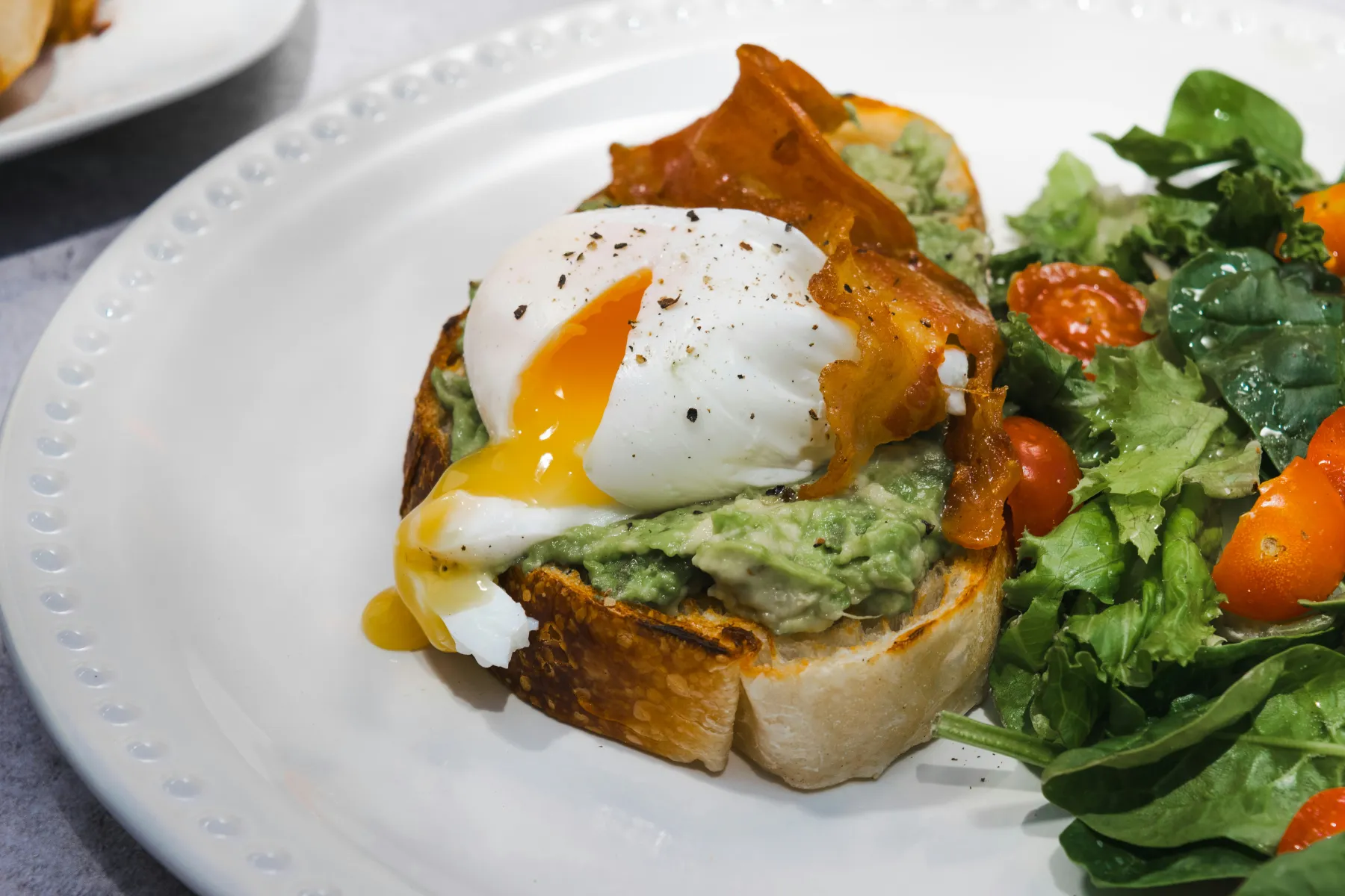 Pocherat ägg Ett pocherat ägg med rinnande gula ligger på avokadotoast med krispig prosciutto och serveras tillsammans med en fräsch sallad med körsbärstomater och bladgrönsaker på en vit tallrik.