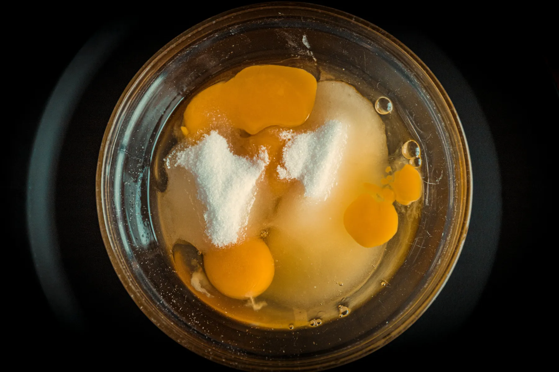 En glasskål med knäckta råa ägg och högar med vitt socker, sedd uppifrån mot en mörk bakgrund.