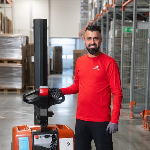 johan-kita-team-ledare En man i röd skjorta och handskar står i ett lager och ler, med ena handen på en orange pallvagn – och tar med självförtroende tag i sin karriär bland hyllor fyllda med lådor och pallar i bakgrunden.