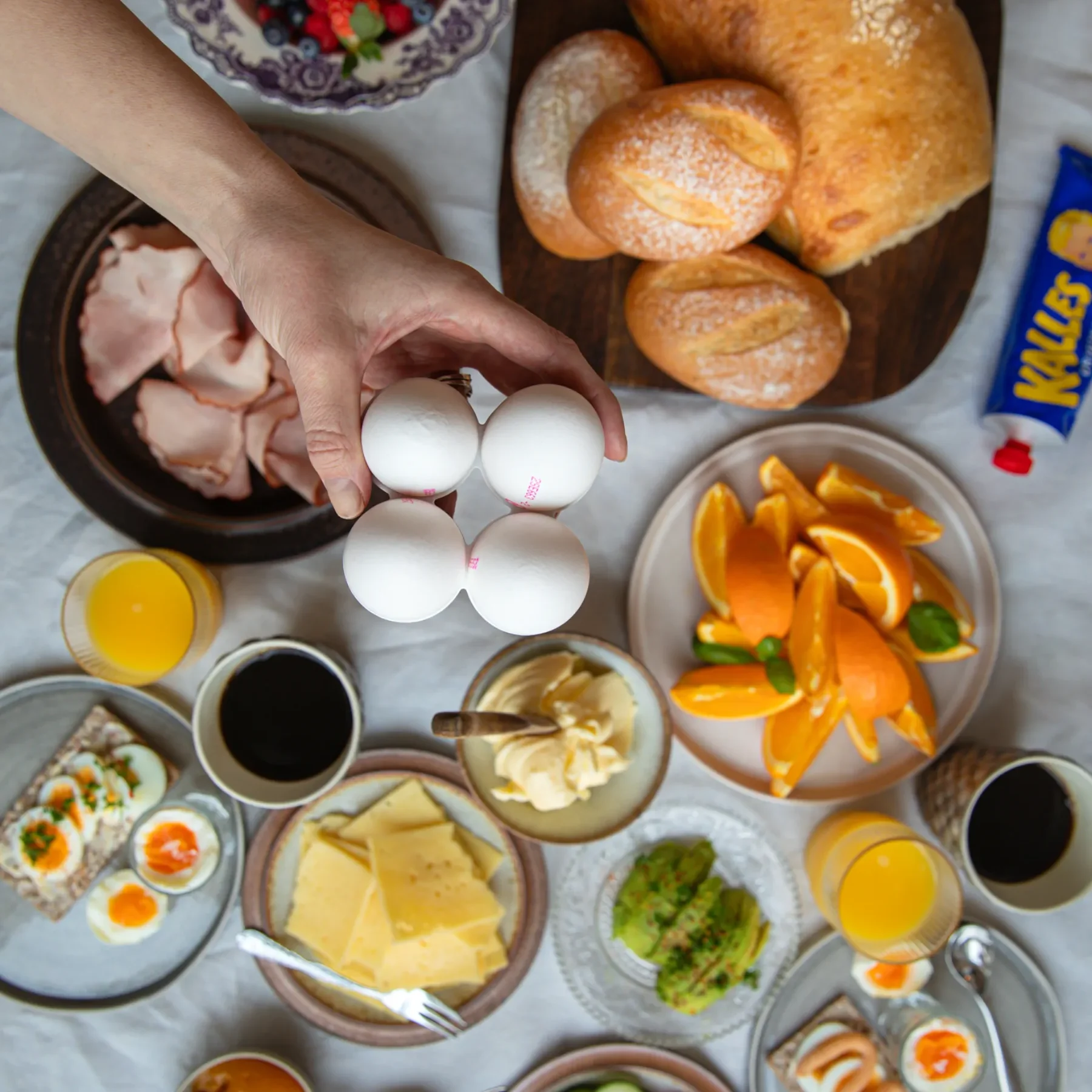 Äggfrukost Ett bord dukat med olika frukosträtter: bröd, frallor, ost, skinka, skivade grönsaker, frukt, apelsinskivor, sylt, avokado, ägg, juice, kaffe och en hand som håller i fyra vita ägg ovanför upplägget.