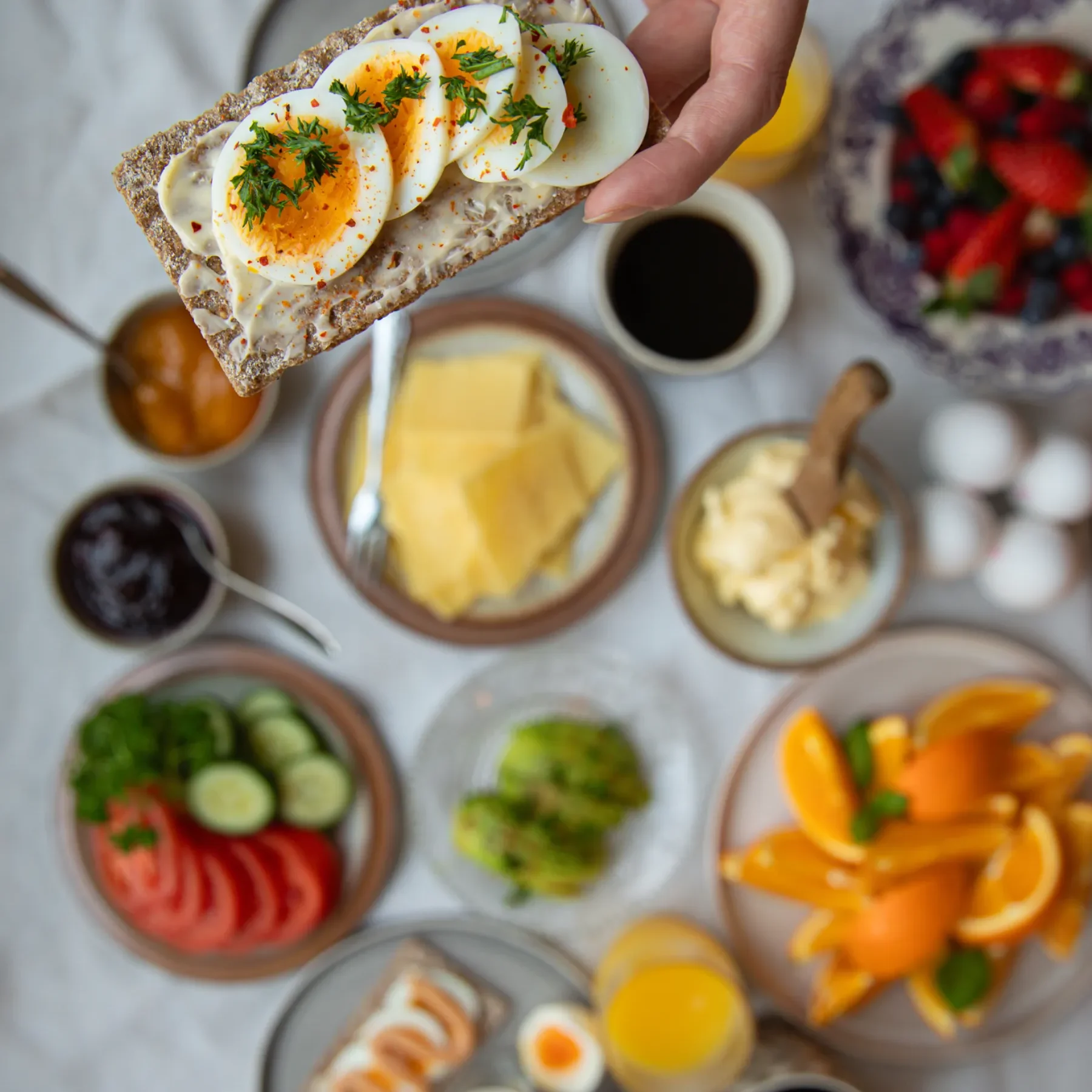 En hand håller i ett knäckebröd med skivade kokta ägg och örter över ett frukostbord med frukt, ost, grönsaker, sylt, smör, apelsinskivor, juice och ägg.