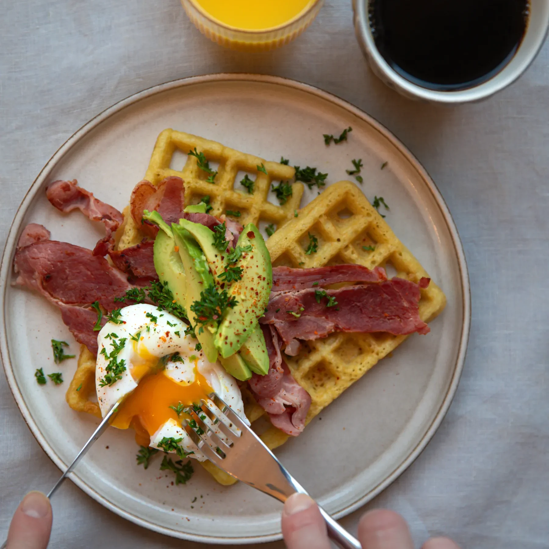 Frukostvåffla En tallrik med våfflor, skivad avokado, knaprigt bacon och ett pocherat ägg som skärs upp med gaffel och kniv. Bredvid tallriken står en kopp apelsinjuice och en mugg svart kaffe på en ljus bordsduk.