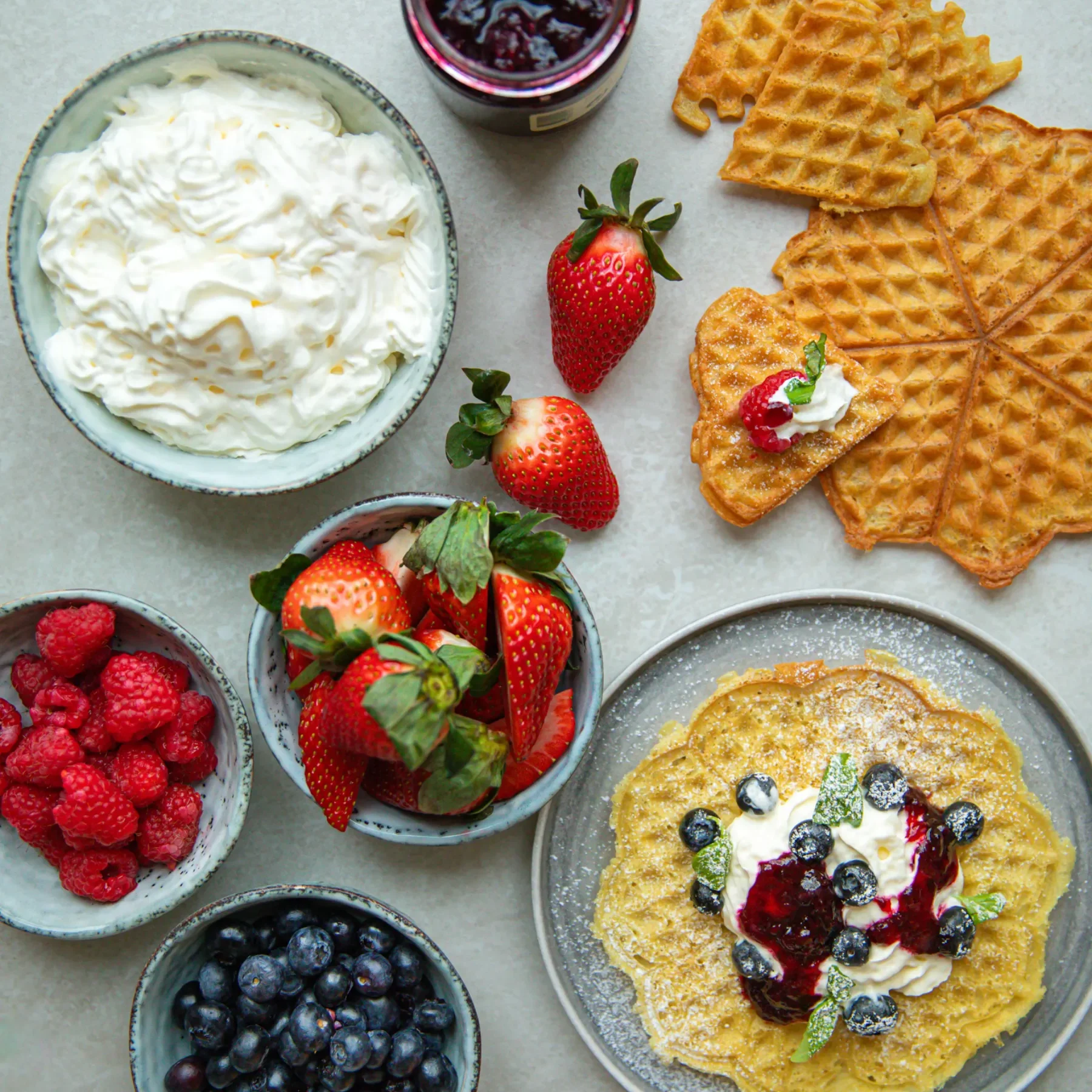 Sommarvåfflor En frukost med våfflor, en skål vispgrädde, en burk bärsylt, färska jordgubbar, hallon, blåbär och en tallrik våfflor toppade med grädde, sylt och bär på en ljus yta.