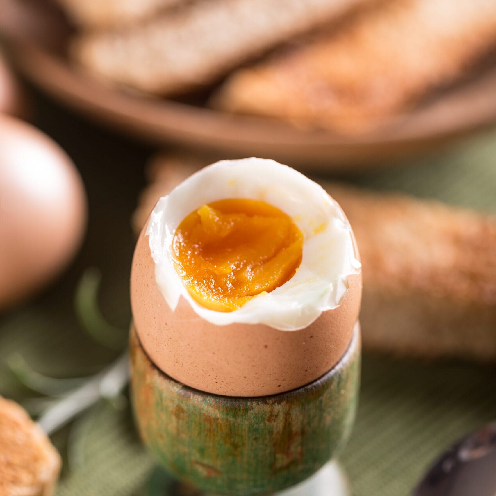 Ett mjukkokt ägg med rinnande gula ligger i en grön äggkopp. Toastsoldater och en sked finns i närheten, och en tallrik med mer rostat bröd är suddig i bakgrunden.